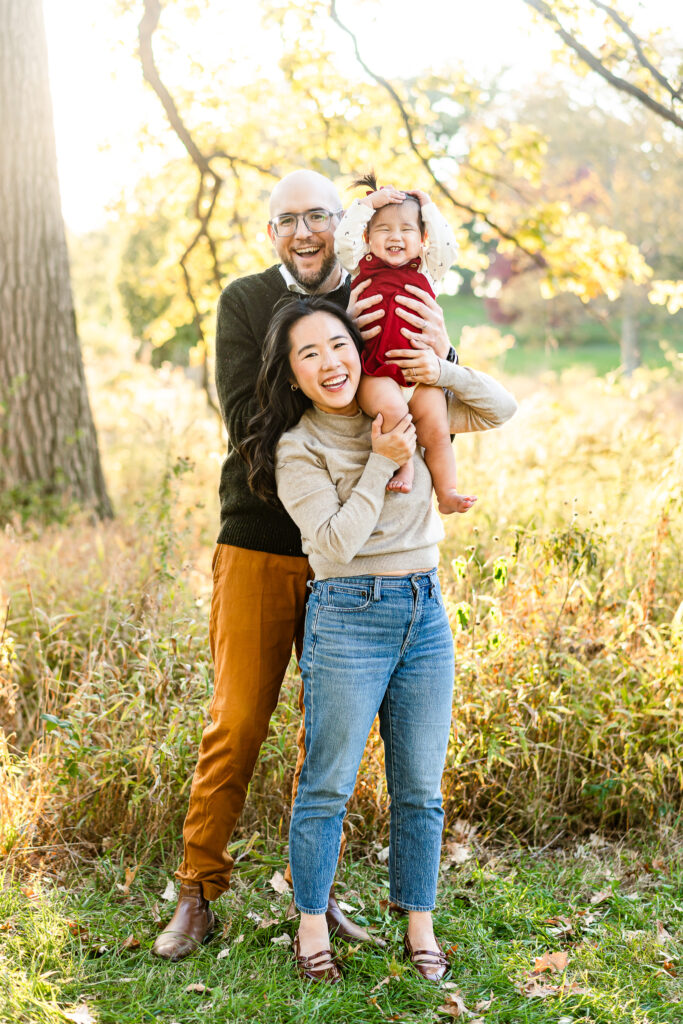 A family smiles happily at the camera. A little girl is held up on her mom's shoulder. 