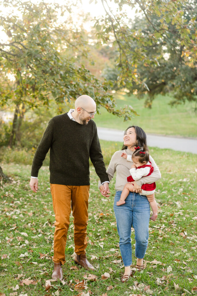 A young mom and dad smiling at each other and holding hands. The mom holds a small girl. 