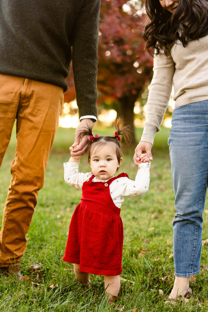A toddler looking at the camera holding her parents hands. 