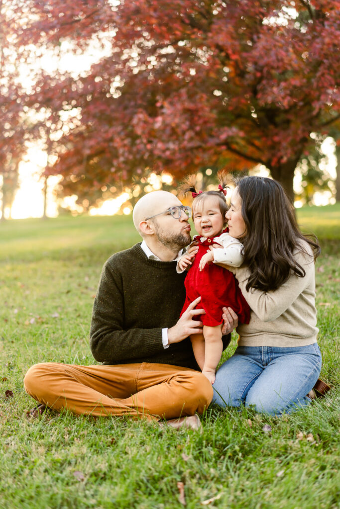 A mom and dad holding their daughter between them leaning in and kissing her cheek in Forest Park.