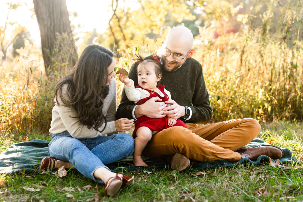 A family sitting on the ground. The little girl hands her mother a leaf she found. 