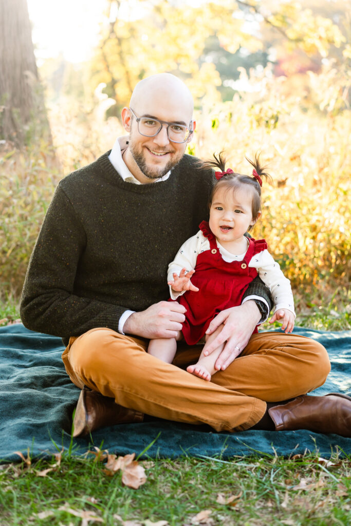 A dad sitting on a blanket with his toddler daughter on his lap. Both are smiling. 
