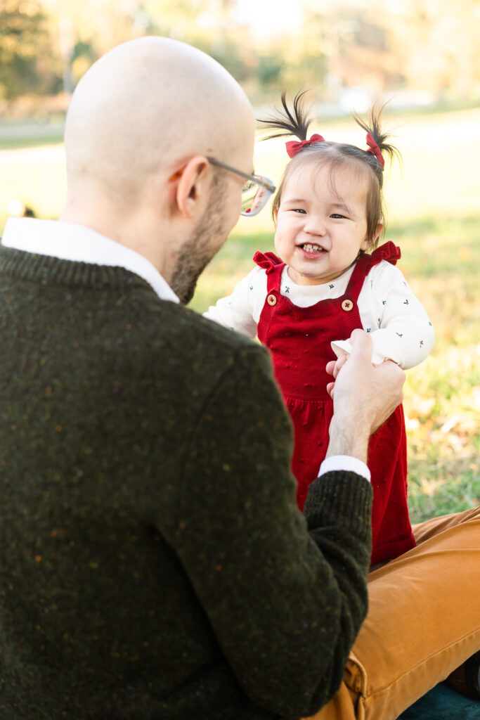 A small girl with pig tails smiling at her father who is holding her hands. 