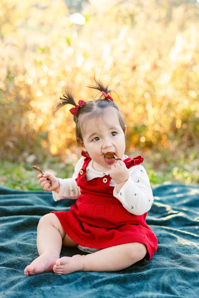 A little girl sits on a blanket trying to eat a leaf. 