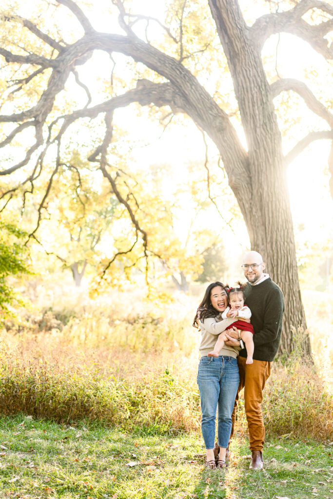 A small family (one woman, one man, and one toddler) smiling at the camera. The mom is holding the toddler and the light is glowing behind them. 