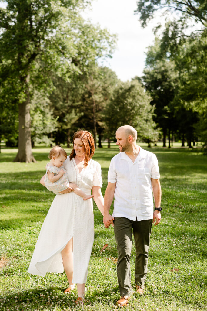 A mom and dad dressed in light colors hold hands and walk towards the camera in Tower Grove Park. The mom holds her young daughter with one arm. Mom and Dad look at their daughter, smiling.