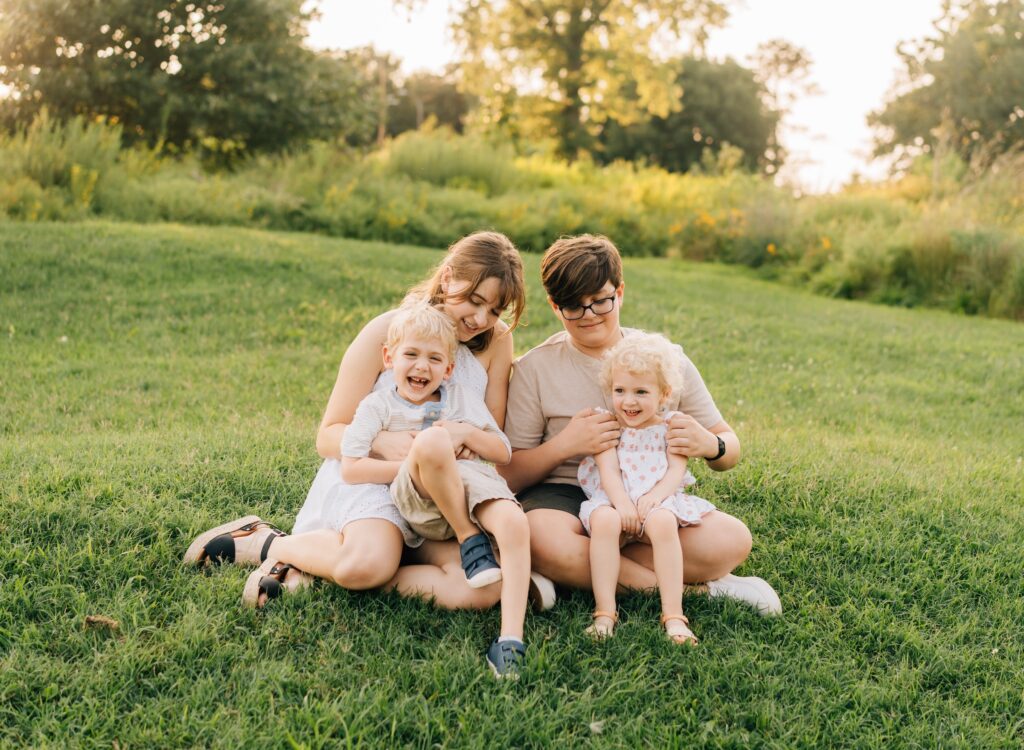 Two older siblings tickle their younger siblings who sit in their laps at the Forest Park Nature Playscape.