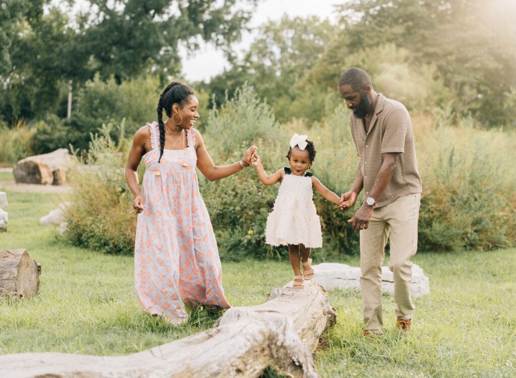 A toddler balances and walks across a log while mom and dad hold her hands. They are smiling.