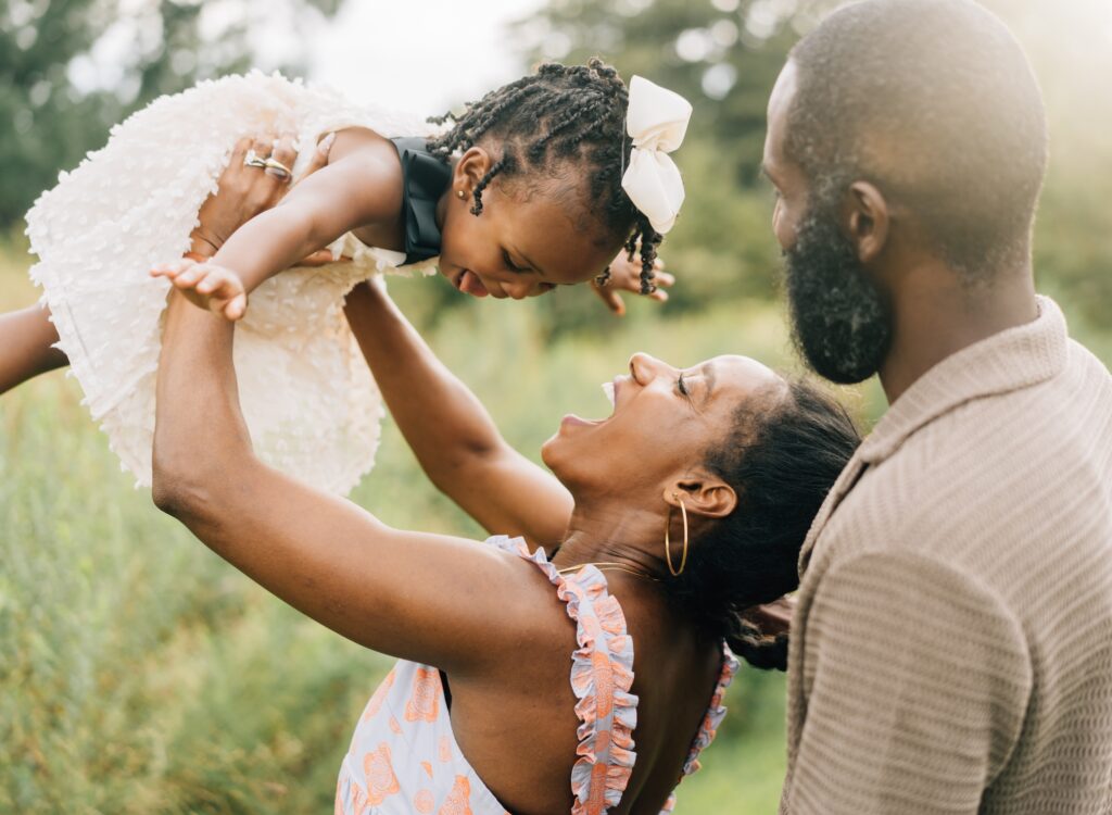 A mom and dad face their daughter and lift her up into the air. She has a big smile on her face.