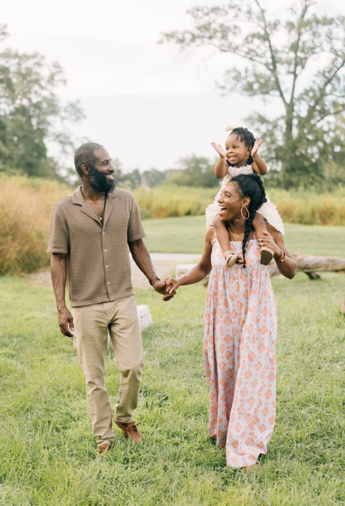 A mom and dad hold hands and the daughter is on the mom's shoulder and they walk and smile at each other.