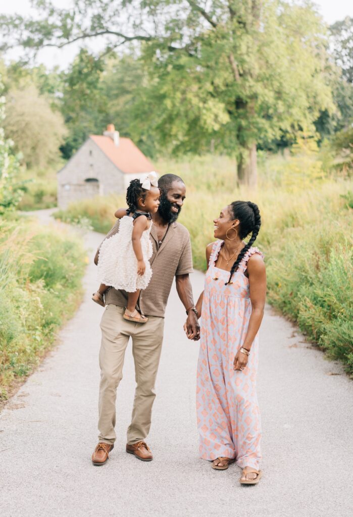 A mom and dad hold hands and smile at each other while the dad holds his little girl. St. Louis outdoor family photos.