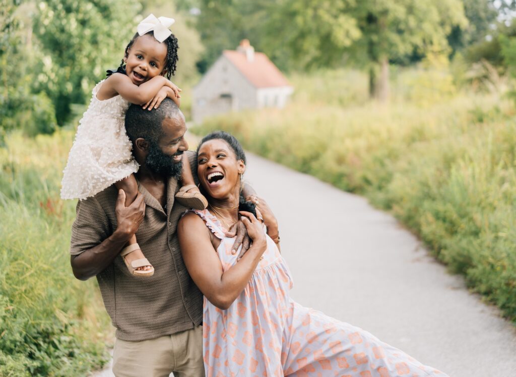 A family of three is laughing and smiling at each other. The mom is leaning back on the dad who has the toddler on his shoulders. They are doing St. Louis Outdoor family photos.