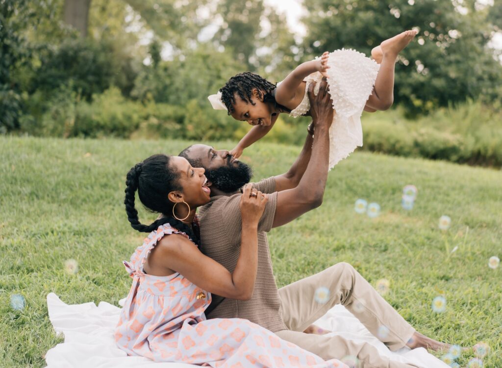A mom and dad sit on a blanket and lift up their daughter above them.