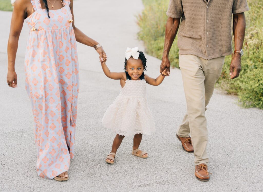 A mom, dad and daughter hold hands and walk. The daughter is looking at the camera and you can only see mom and dad's bodies.