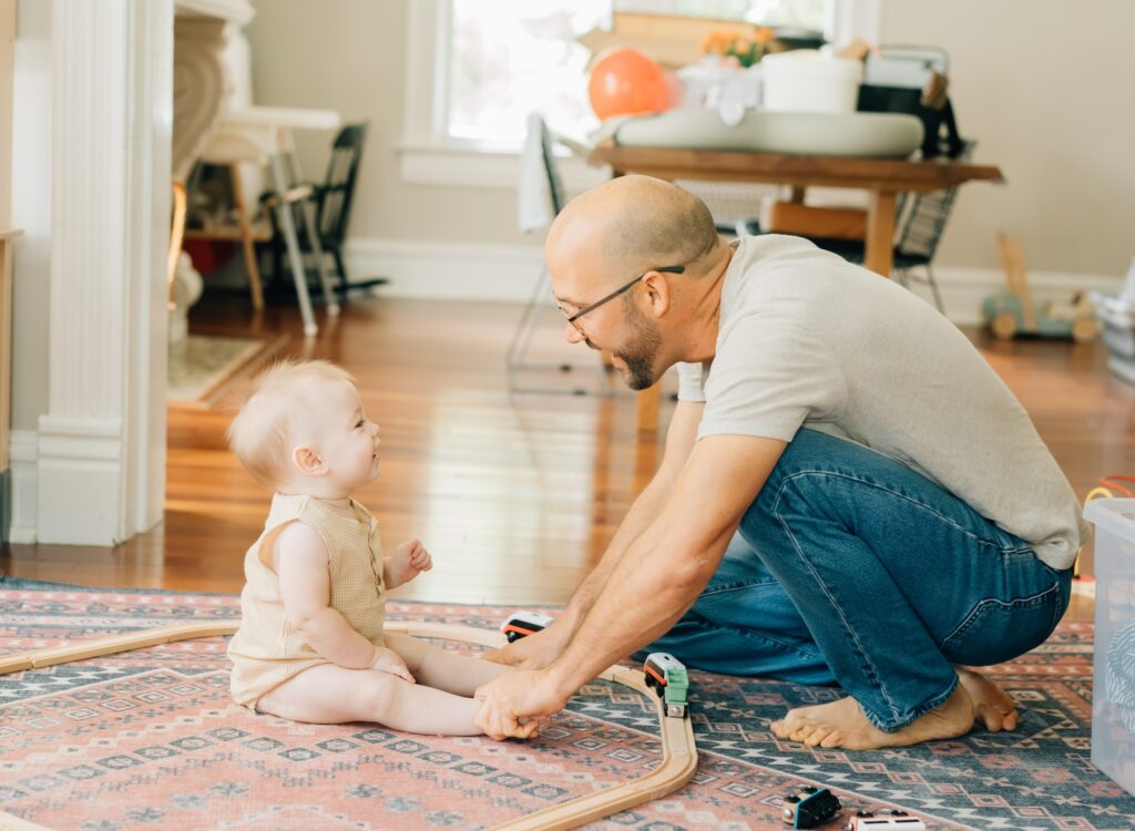 A father crouched down and holding his little ones feet. They are smiling at each other.
