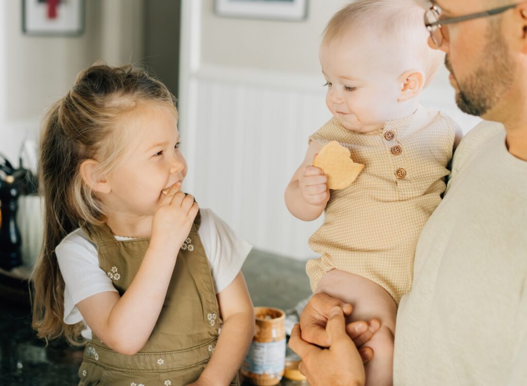 A toddler and baby snacking on some crackers and looking at each other.