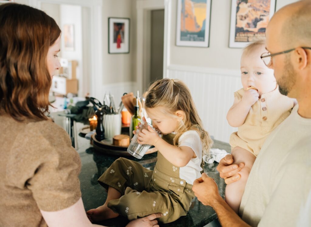 A girl sitting on the counter drinking water while a baby snacks on a cracker and the parents watch them.
